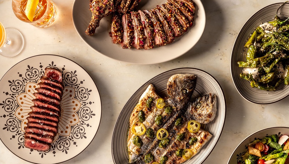 Restaurant table featuring sliced steak, grilled whole fish, fresh salads, roasted vegetables, and cocktails served family-style